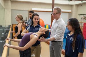 A faculty member in a white shirt is holding the back of the head of a ballet dancer as they lean back. Two students in blue shirts stand on either side observing