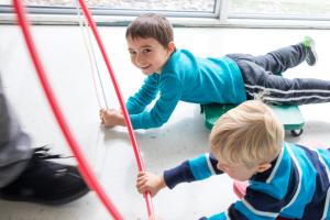 Two children are on scooters being pulled down a hallway with a hulahoop.